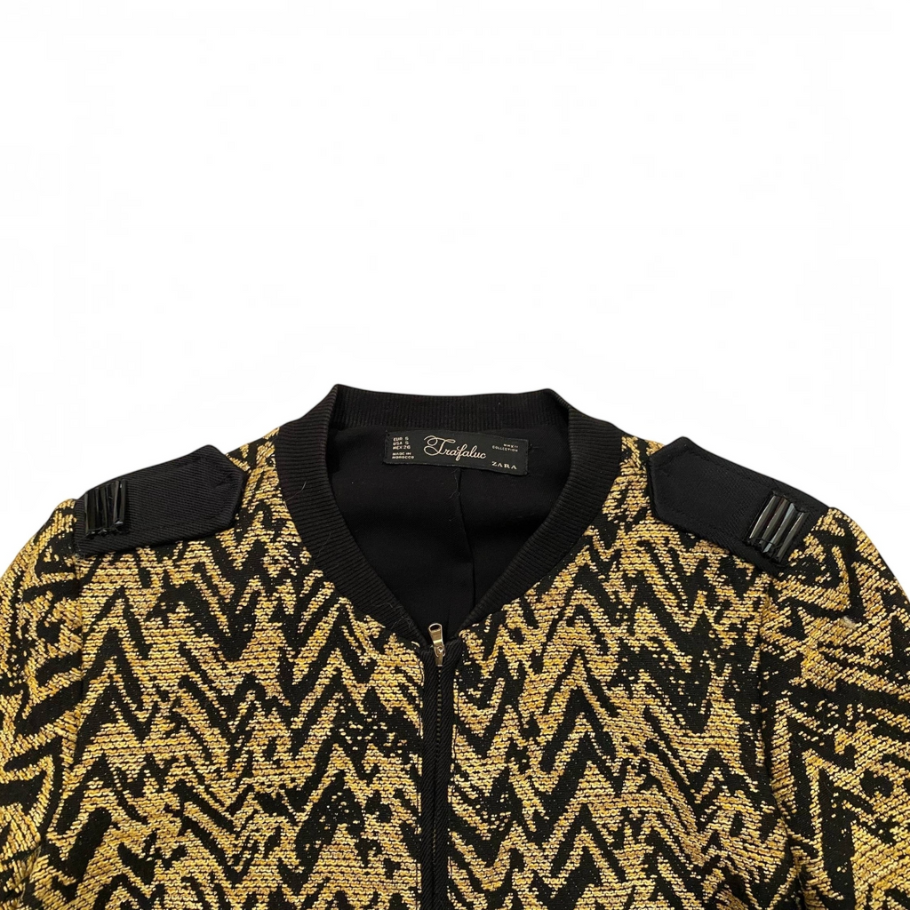 Chaqueta Zara Trafaluc con estampado en zigzag negro y dorado, cierre de cremallera y detalles en hombros.
